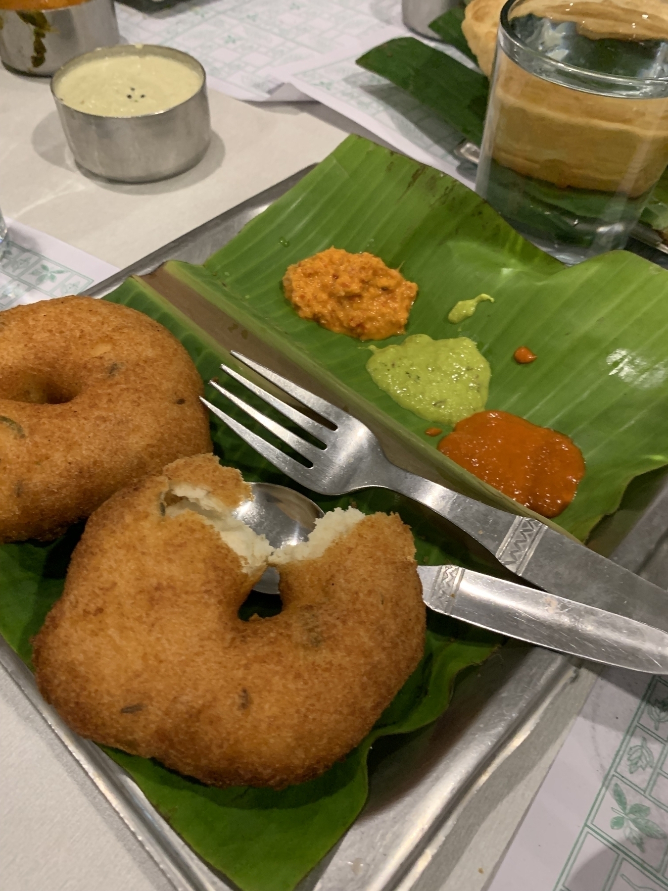 Two vadas are served on a banana leaf along with three types of chutneys and a glass of water in the background.