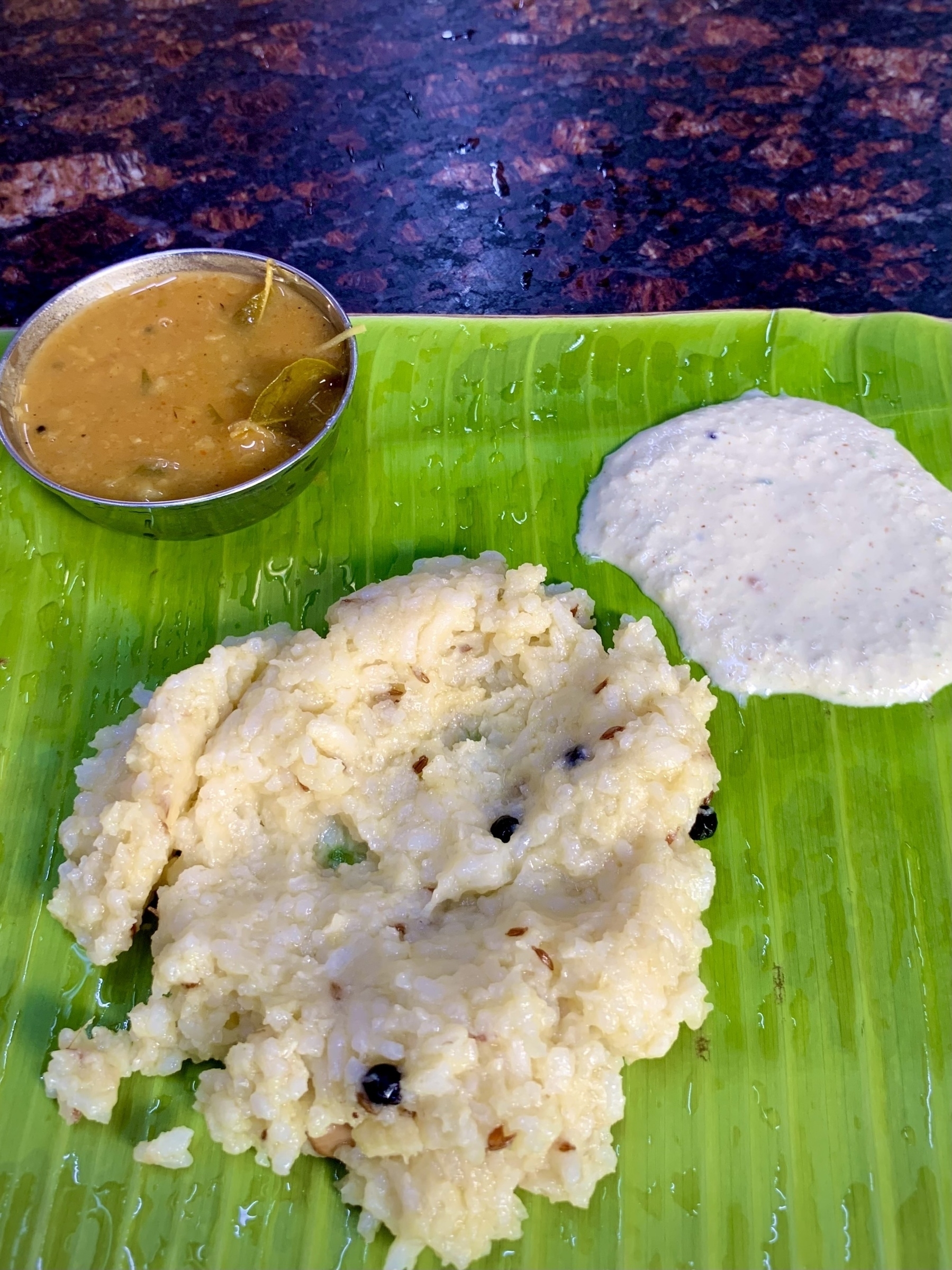 A serving of ghee pongal, sambar in a small bowl, and coconut chutney is presented on a banana leaf.