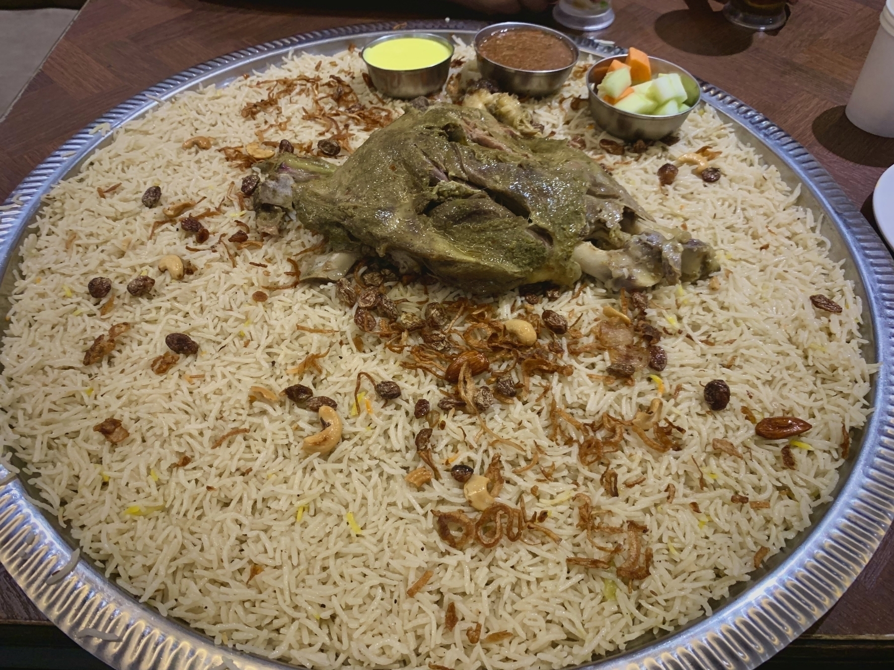 A large platter of Mandi is topped with a piece of cooked lamb leg, garnished with fried onions and nuts, and accompanied by small bowls of sauces and vegetables.