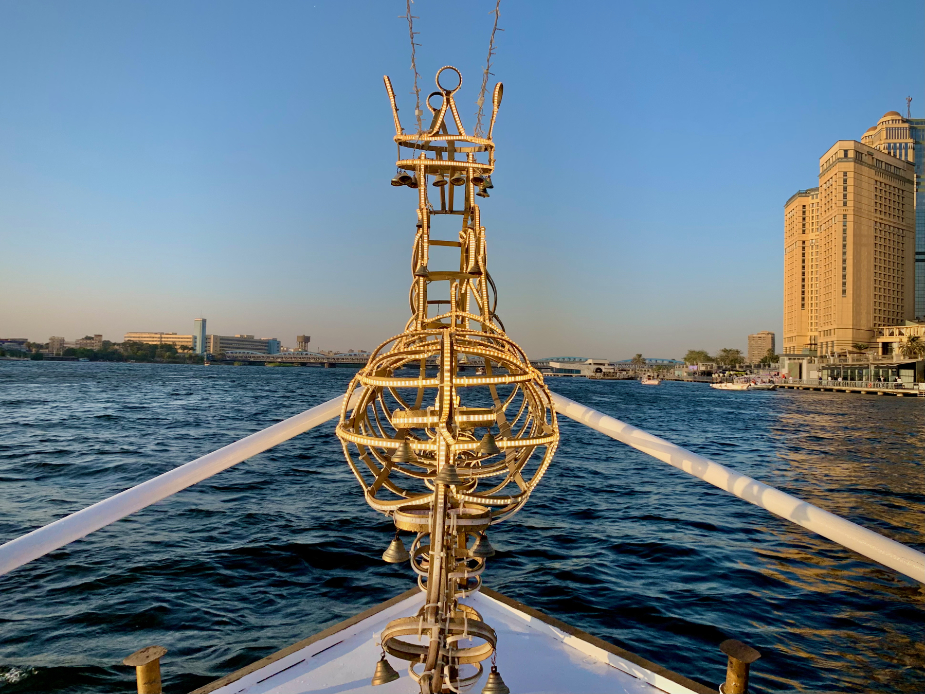 Auto-generated description: A boat travels along a river with a decorative metal structure at its bow and city buildings in the background.