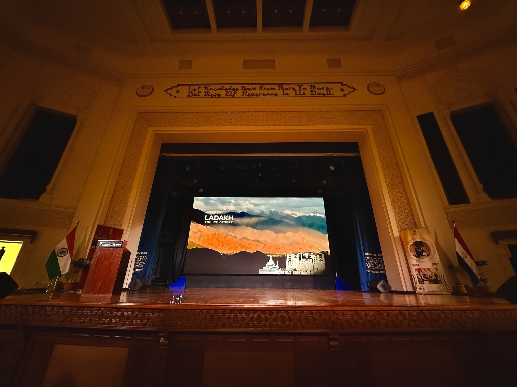A stage is set up with a screen displaying a scenic image of Ladakh, flanked by flags and a podium in a decorated hall.
