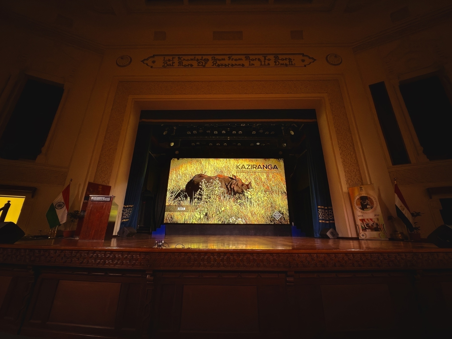 A stage with a large screen displays an image of a rhinoceros in a grassy field, alongside the text Kaziranga, with flags and banners nearby.