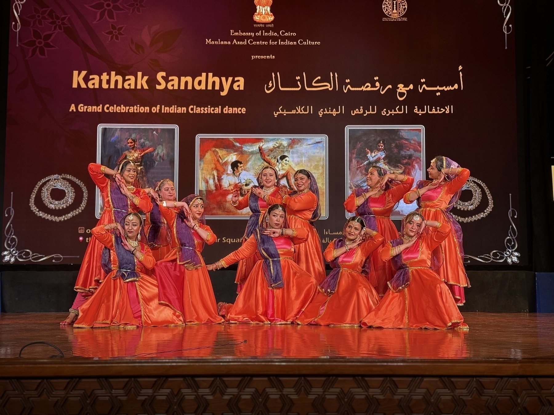 A group of dancers in vibrant orange and red costumes perform on stage against a backdrop promoting a classical dance event.