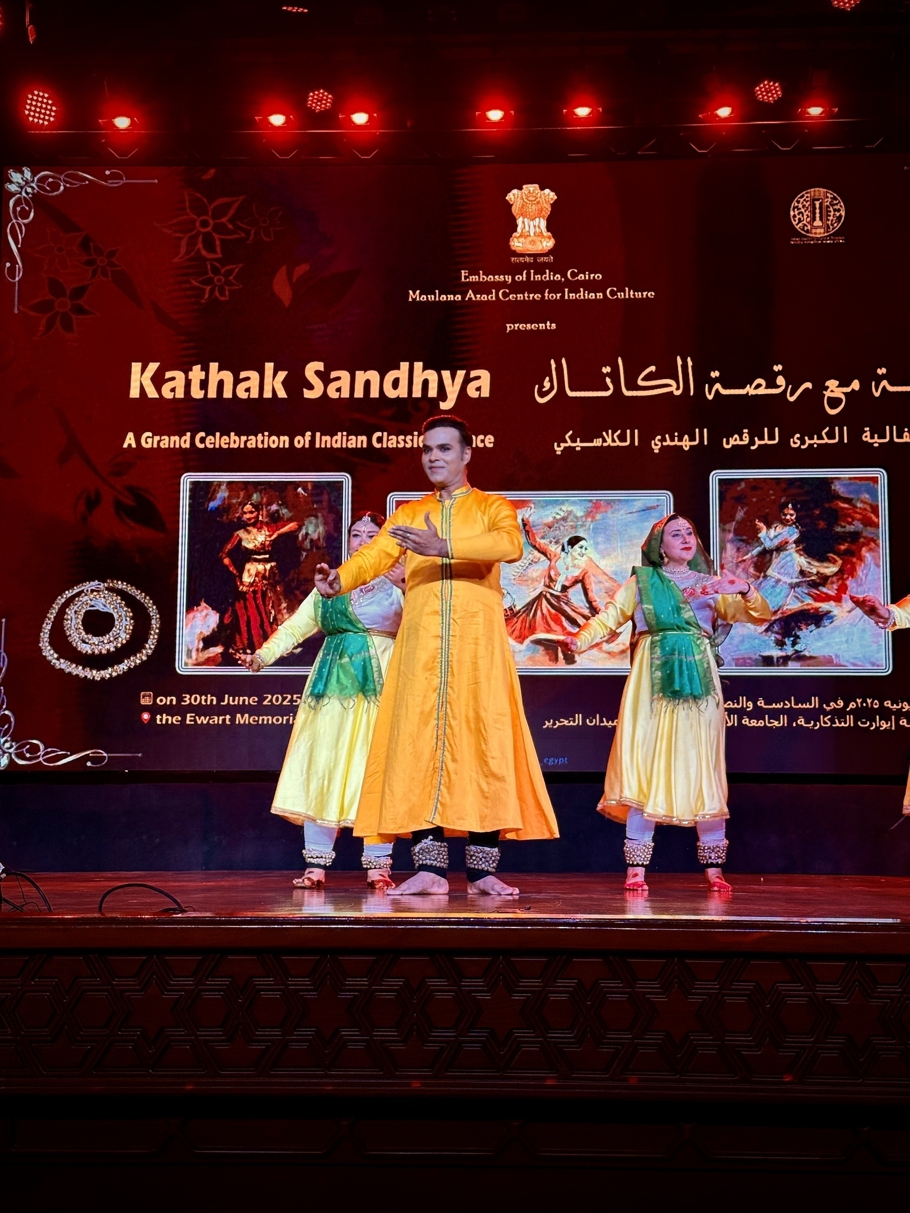 A group of performers in colorful traditional attire is dancing on stage under vibrant lighting during an event named Kathak Sandhya.
