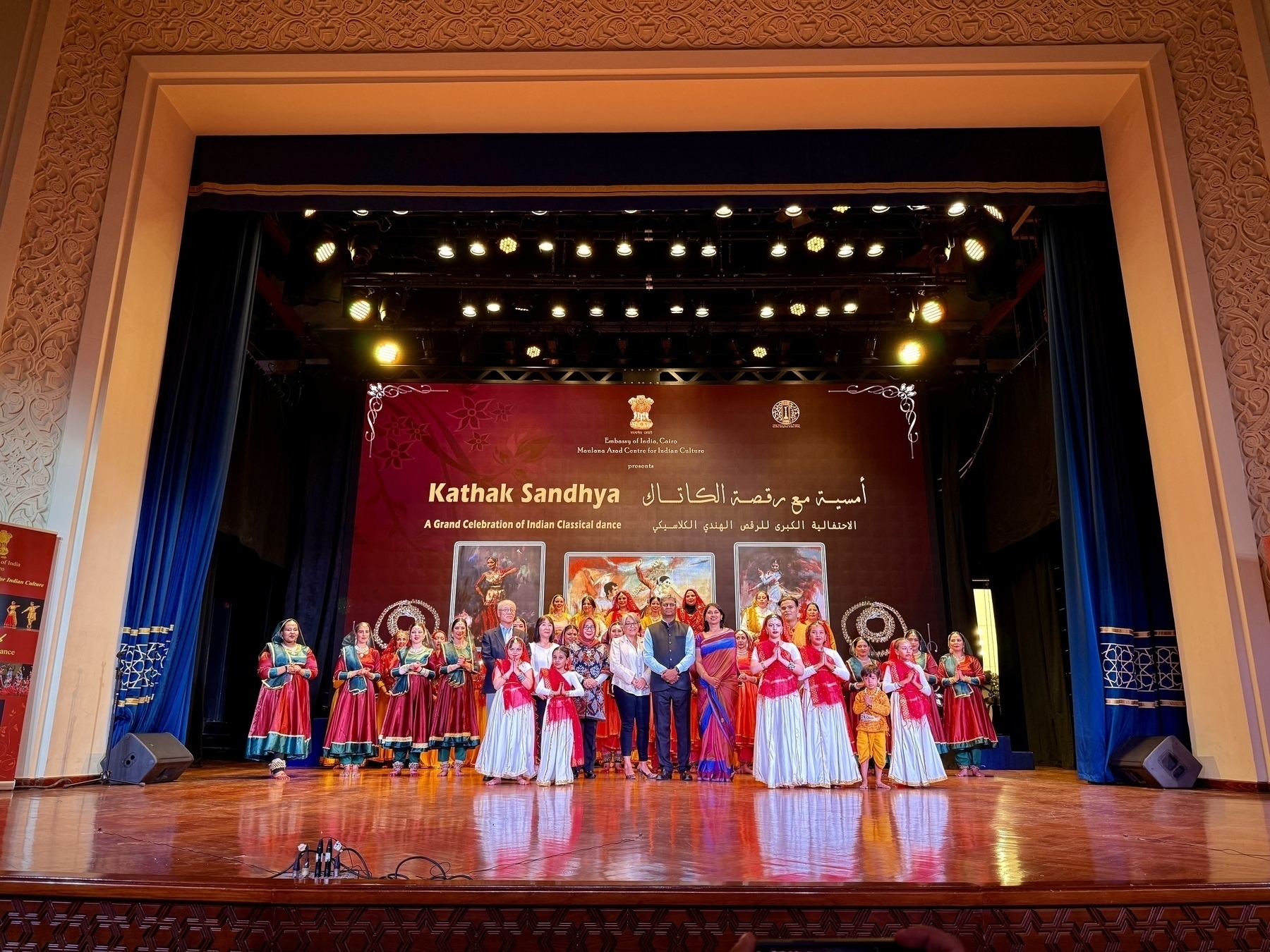 A group of performers in traditional attire pose on stage during the Kathak Sandhya event, surrounded by ornate decorations and lighting.