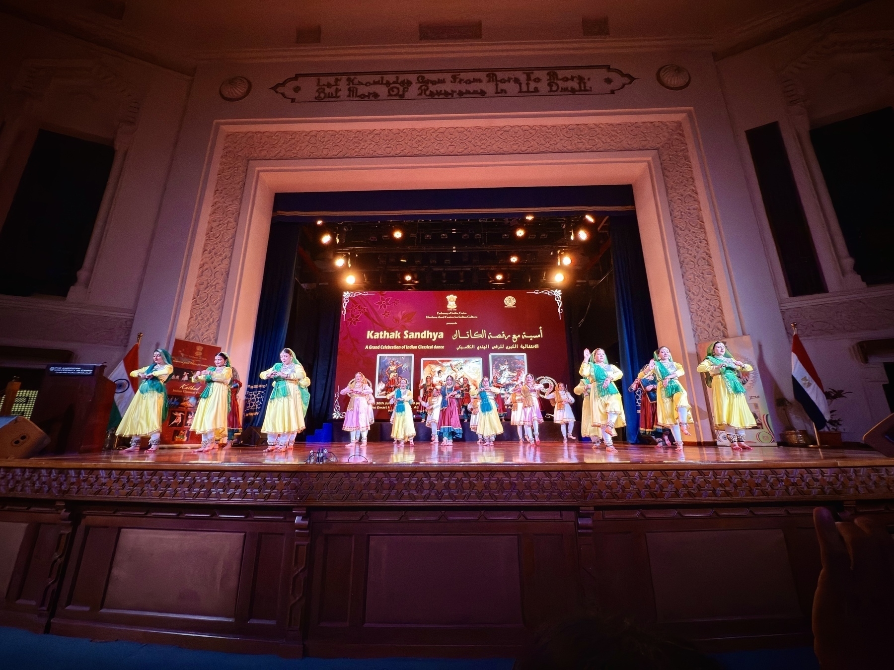A group of performers in colorful traditional costumes are dancing on a well-lit stage with an ornate, decorated backdrop.