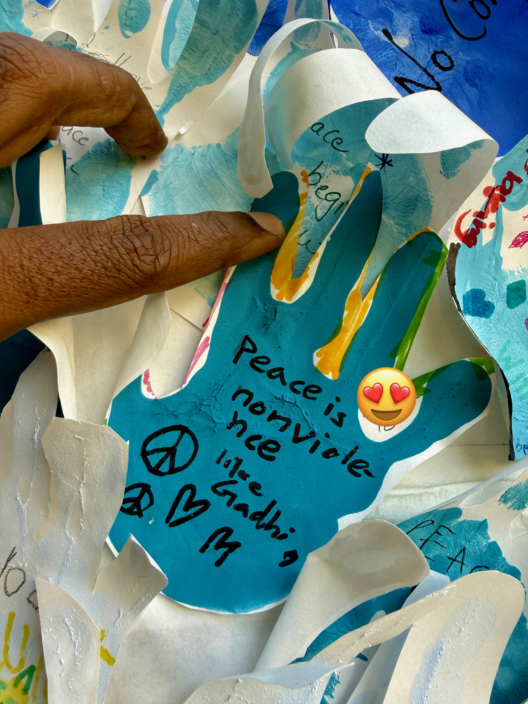 A hand-shaped paper cutout with messages about peace and love is surrounded by similar blue cutouts, with a person's fingers touching one.