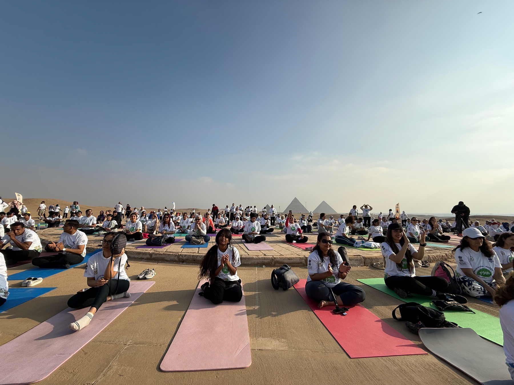 A large group of people are practicing yoga on mats near the Great Pyramids of Giza under a clear sky.