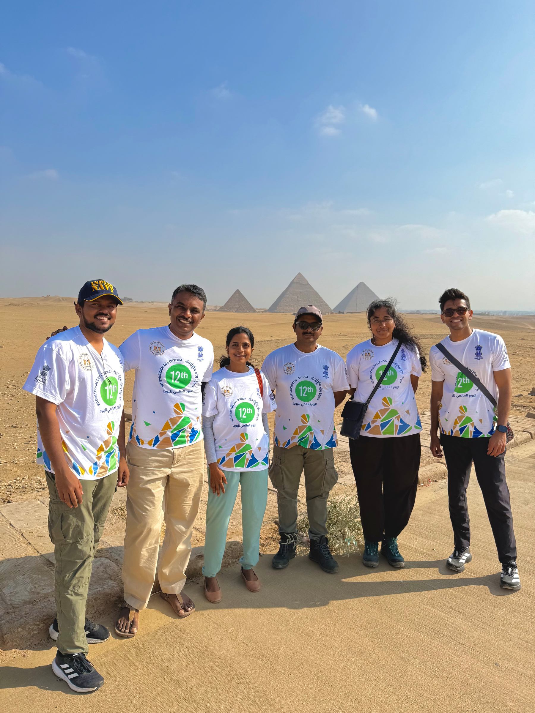 A group of six people wearing matching white shirts with colorful designs are standing in front of the pyramids under a clear blue sky.