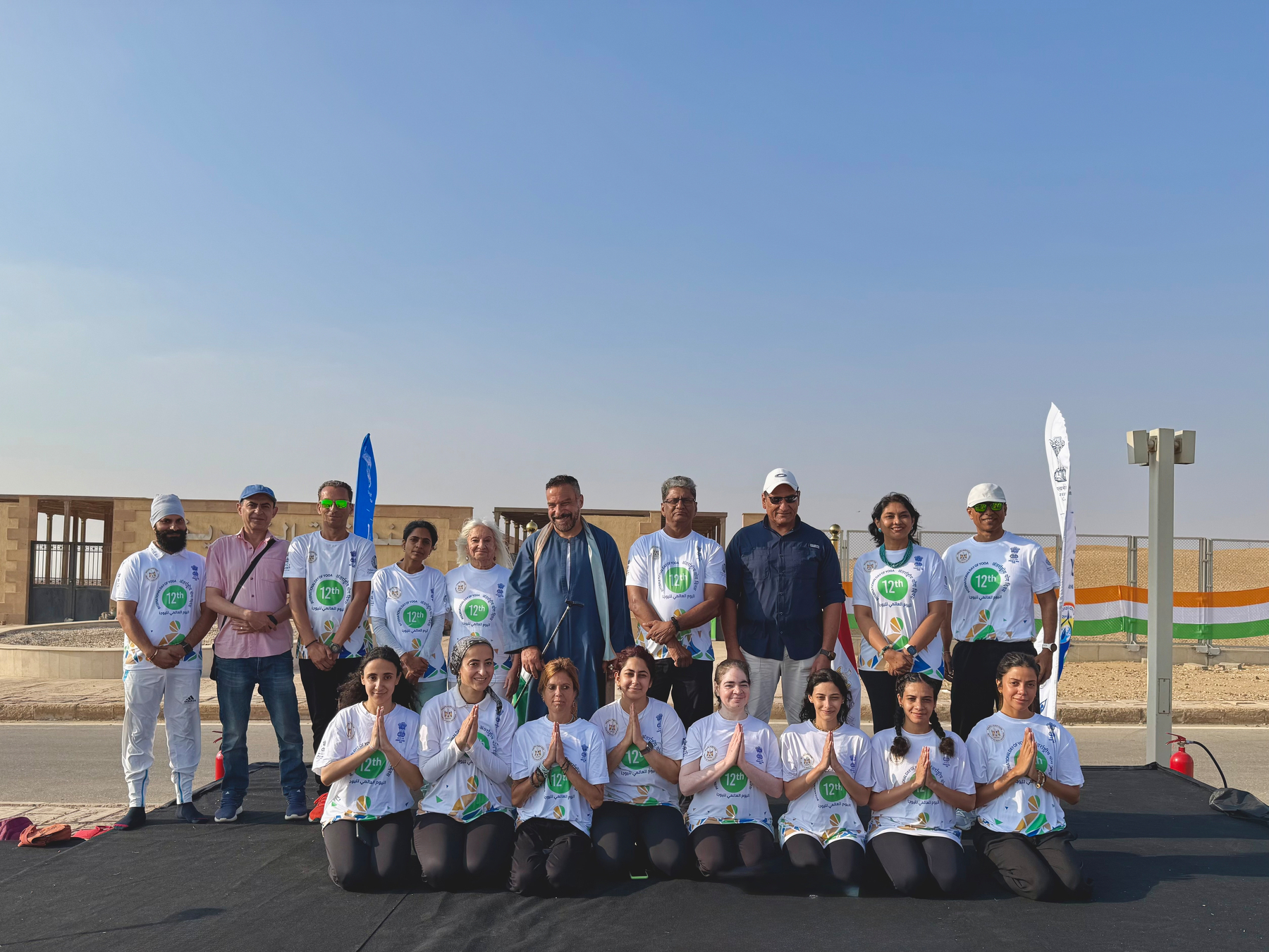 A group of people, some standing and some kneeling, are posing outdoors wearing matching white shirts with green and blue logos, with a clear sky and a desert-like background.
